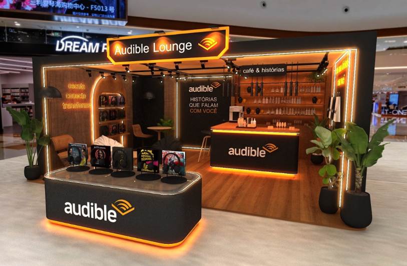 Espaço Audible Lounge em shopping, com balcão, assentos, exposição de títulos e iluminação laranja.